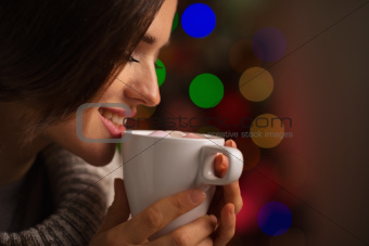 Happy young woman enjoying cup of hot beverage in front of Christmas lights