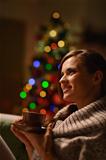 Thoughtful woman sitting chair and drinking hot beverage in front of Christmas tree