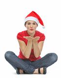 Happy young woman with Christmas hat sitting on floor and blowing kiss