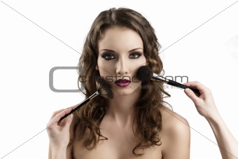 girl looks in to the lens, getting made-up by hands with brushes