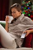 Young woman drinking hot chocolate and watching TV in front of Christmas tree