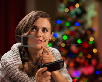 Thoughtful young woman writing sms in front of Christmas tree