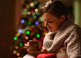 Dreaming young woman sitting chair with hot chocolate in front of Christmas tree