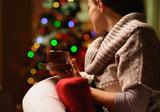 Young woman sitting chair with hot beverage in front of Christmas tree. Rear view
