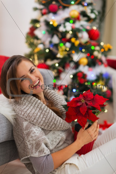 Smiling woman with Christmas rose sitting near Christmas tree
