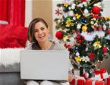 Smiling young woman with laptop sitting near Christmas tree