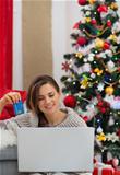 Smiling woman with laptop and credit card sitting near Christmas tree