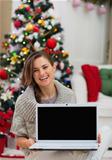 Smiling woman showing laptop blank screen near Christmas tree
