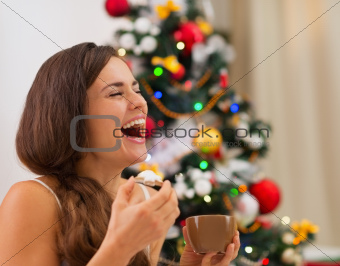 Smiling young woman in pajamas eating cookies with hot chocolate near Christmas tree