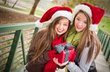 Two Attractive Festive Smiling Mixed Race Women Wearing Christmas Santa Hats Holding a Wrapped Gift with Bow Outside.