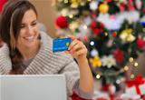 Closeup on credit card in hand of happy woman with laptop near Christmas tree