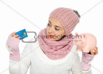 Confused woman in knit winter clothing holding credit card and piggy bank