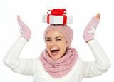 Happy woman in knit winter clothing balancing Christmas present box on head