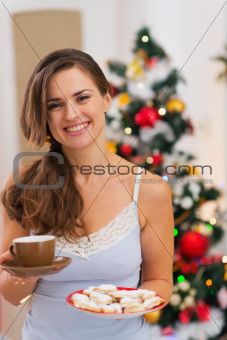 Happy woman in pajamas holding hot beverage and cookies in front of Christmas tree