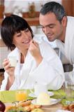a woman giving a teaspoonful of yoghurt to his husband at breakfast, both are wearing bathrobes