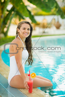 Smiling young woman sitting on pool edge with cocktail