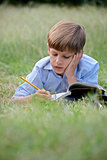 Young school boy doing homework alone, lying on grass