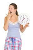 Young woman in pajamas with clock yawing after sleep