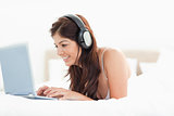 Woman browsing her laptop with headphones while lying on a bed