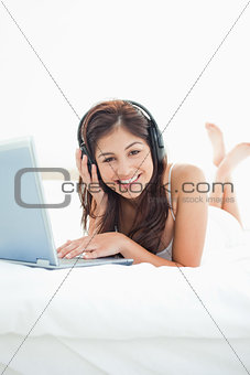 Woman with headphones and a laptop lying on bed, looking forward