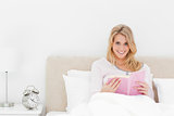Woman in bed reading a book, while looking forward and smiling