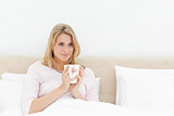 Woman in bed with a cup in hands, softly smiling while looking t