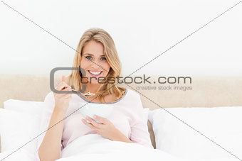 Woman in bed, holding a bowl and spoon of cereal looking forward