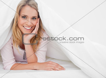 Woman lying in bed under quilt raised to her head, with her hand
