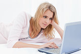 Woman lying on bed scrolling on her laptop while resting her hea