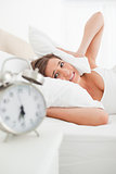 Woman covers her ears with a pillow and looks at her clock in an