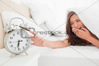 Woman yawning and reaching out for alarm clock from under her bl