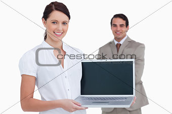 Smiling saleswoman presenting laptop screen with colleague behin