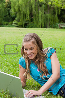 Young smiling girl lying down in a park while using her laptop