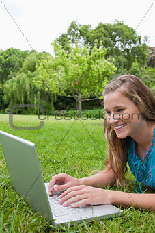 Young smiling girl using her laptop in a park while lying on the