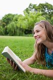 Young smiling woman lying in a parkland while holding a book