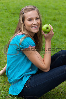 Smiling young woman holding a green apple while sitting in a par