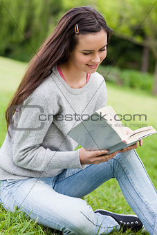 Young relaxed girl sitting on the grass in a park while reading 