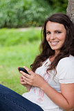 Young smiling woman sitting against a tree while sending a text