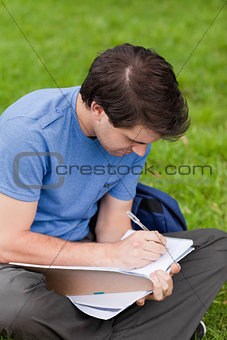 Young student sitting on the grass while writing on his notebook