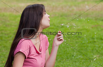 Young woman standing up in a park while blowing a dandelion