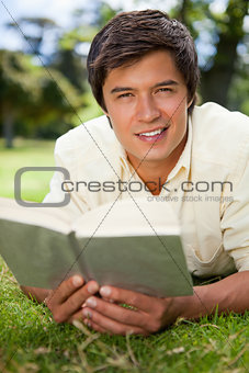 Man looking ahead while reading a book as he lies on grass
