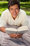 Man smiling while reading a book as he lies on a blanket