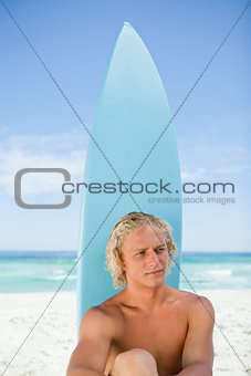 Serious blonde man sitting down in front of his surfboard