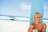 Blonde man sitting in front of his surfboard while looking towar