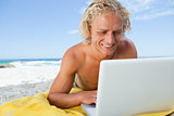Smiling blonde man looking at his laptop while lying on the beac