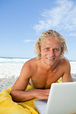 Smiling blonde man looking at the camera while using his laptop
