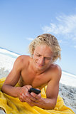 Blonde man lying on his beach towel while sending a text