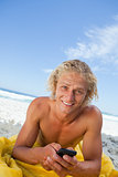 Smiling blonde man sending a text while sunbathing on the beach