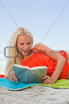 Young smiling woman lying on her beach towel while reading a book
