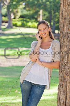 Smiling woman on her cellphone leaning against a tree